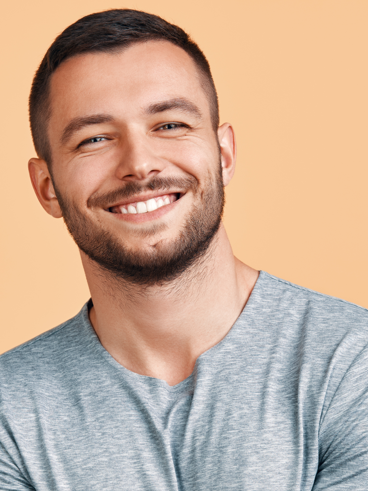 A smiling man with short dark hair and a beard, wearing a gray shirt against a peach background, exudes vitality reminiscent of NAD+ Injections.