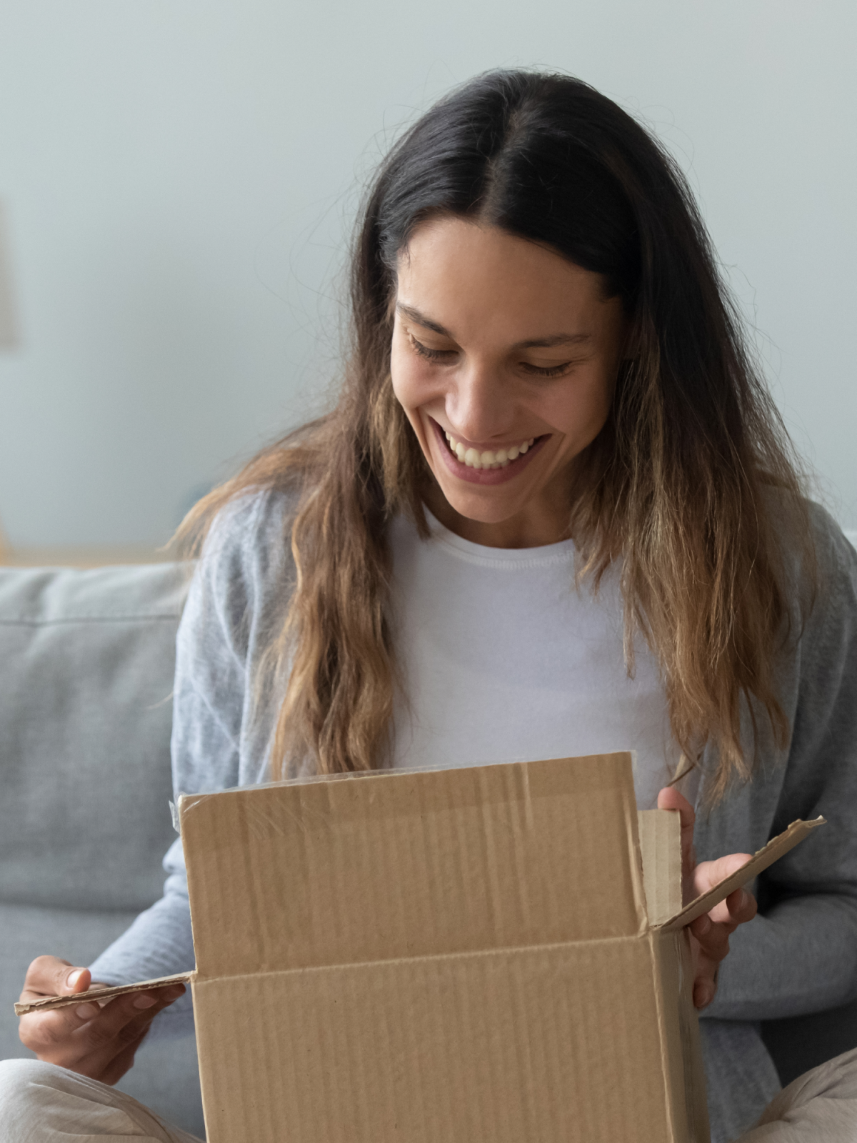 A woman eagerly opens a cardboard box on her couch, smiling at the NAD+ Injections inside, which promise to revitalize her daily routine.