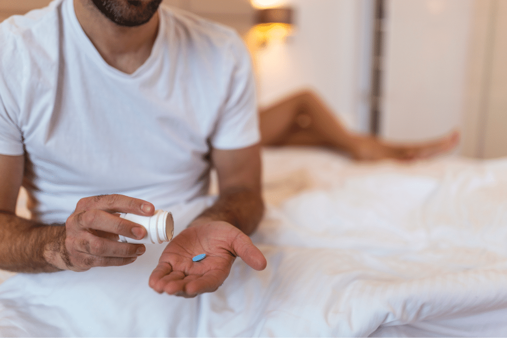 A man holds a pill, either scream cream or Viagra, in his hand while sitting on a bed.