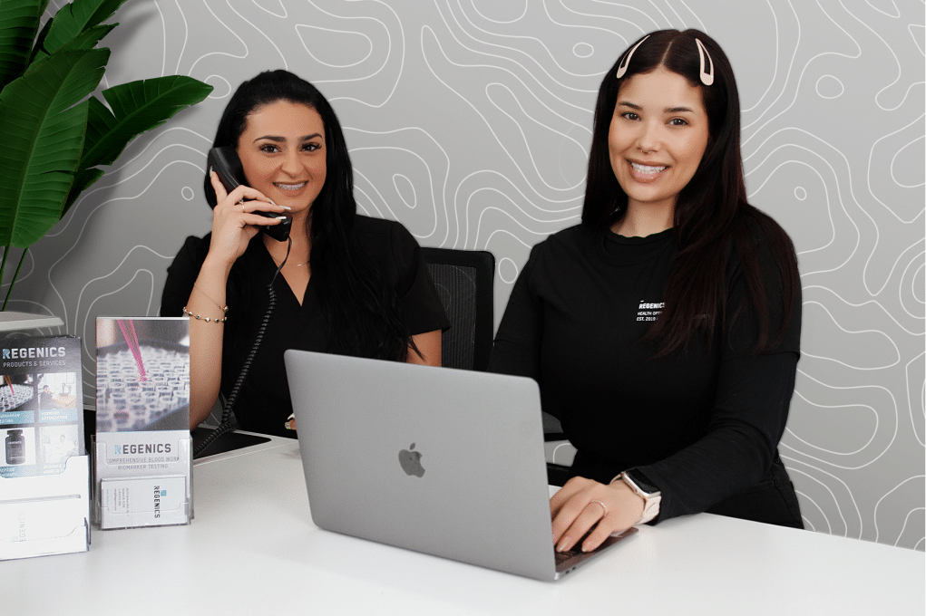 Two women sitting at a desk with a laptop in front of them, engaged in telehealth.