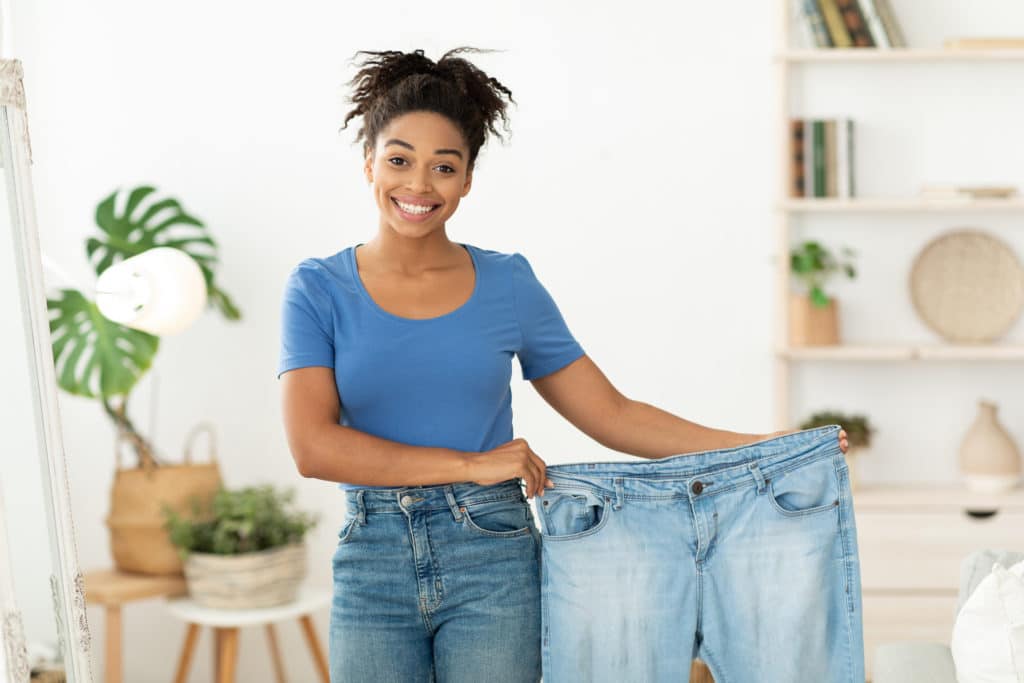 Weight Loss. Happy Slim Black Girl Showing Old Oversize Jeans After Successful Diet Slimming Standing At Home