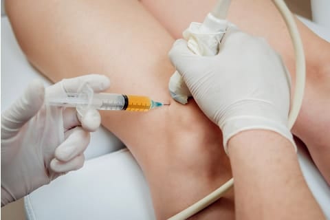 A healthcare professional wearing gloves administers an injection into a patient's knee, using a syringe and ultrasound guidance.