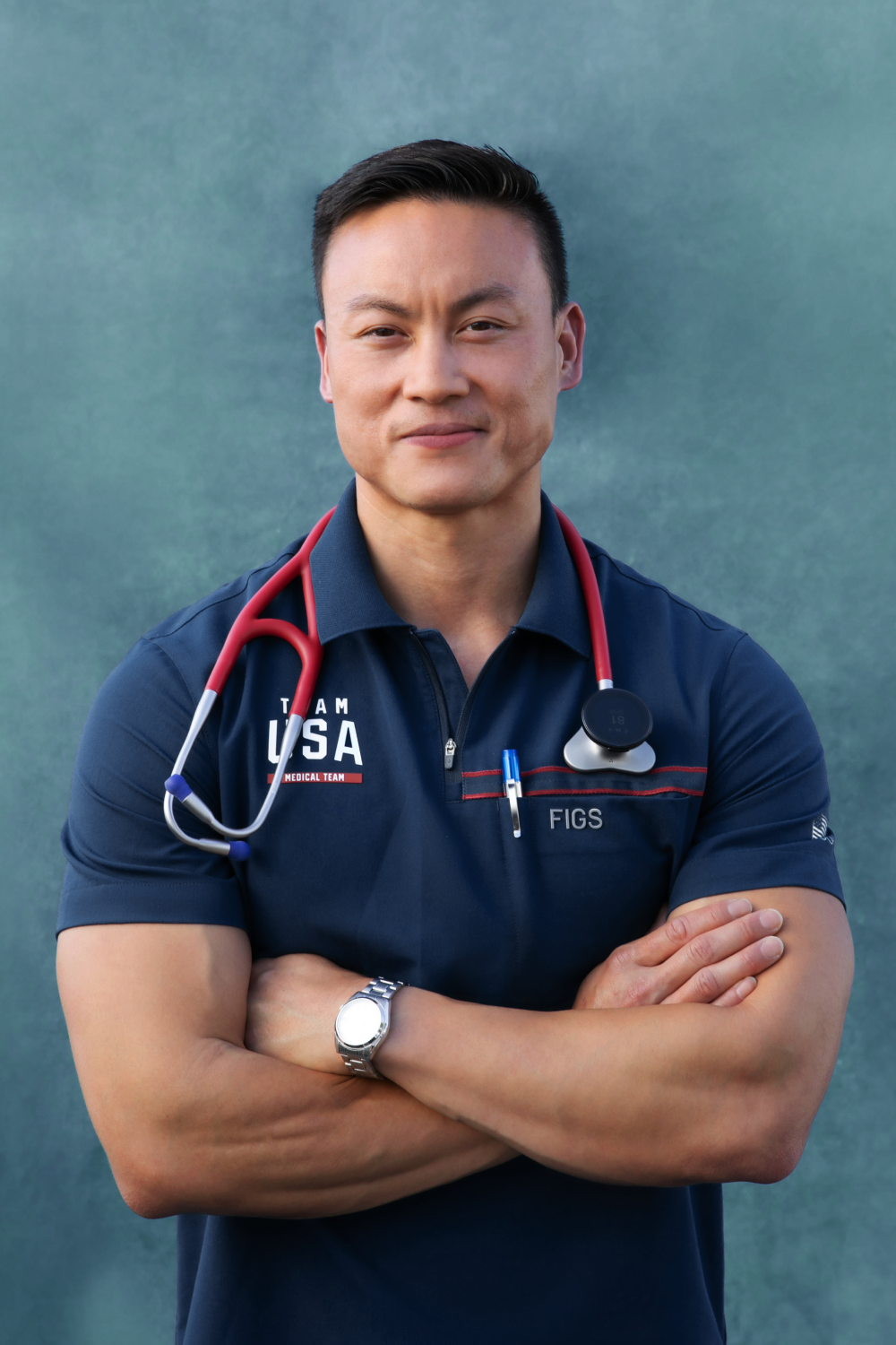 A person donning a navy blue "Team USA" shirt and a red stethoscope around their neck stands confidently with crossed arms against a blue backdrop, embodying the readiness of Executive Physicals.