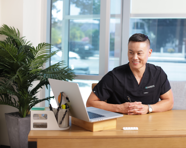 A person in a black medical uniform sits at a desk, immersed in the Prime data displayed on the laptop screen. A window and a potted plant provide a serene backdrop, perfect for delving into the latest Regenics research.