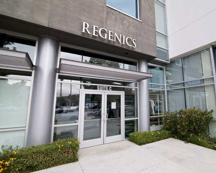 The entrance to the Regenics building showcases sleek glass doors and windows, framed by a "Suite C" sign. Prime landscaping with bushes adorned in yellow flowers lines the walkway, adding a touch of nature's elegance to the scene.