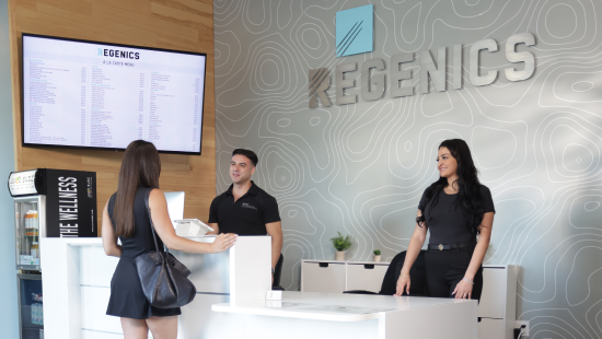 Two employees greet a customer at the reception desk with "Regenics Prime" signage behind them. A large screen displays information.