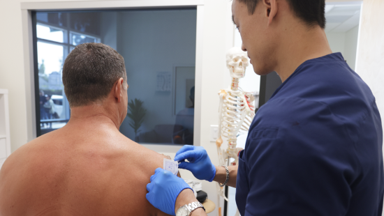 In a clinic room adorned with a skeleton model, a healthcare professional from Regenics, wearing gloves, carefully prepares to inject a man's shoulder.