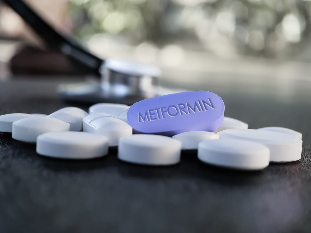 A purple Metformin pill surrounded by white pills on a dark surface, with a stethoscope blurred in the background