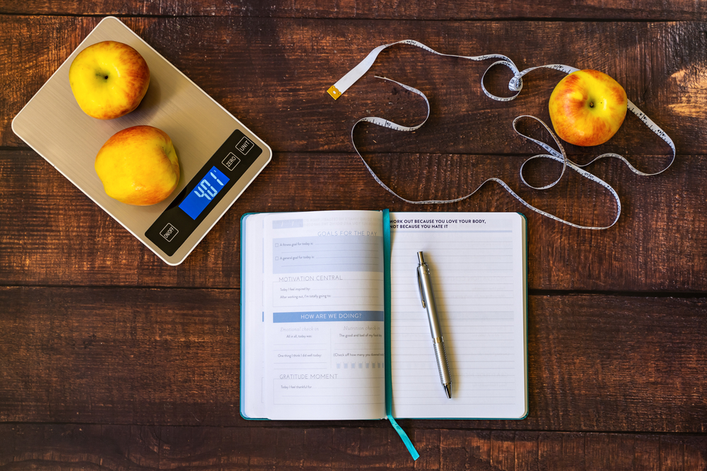 Electronic scales, apples, workout log book and measuring tape on a wooden table