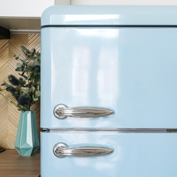 A vintage-style light blue refrigerator with chrome handles stands next to a teal vase holding dark green foliage on a wooden counter in the kitchen.