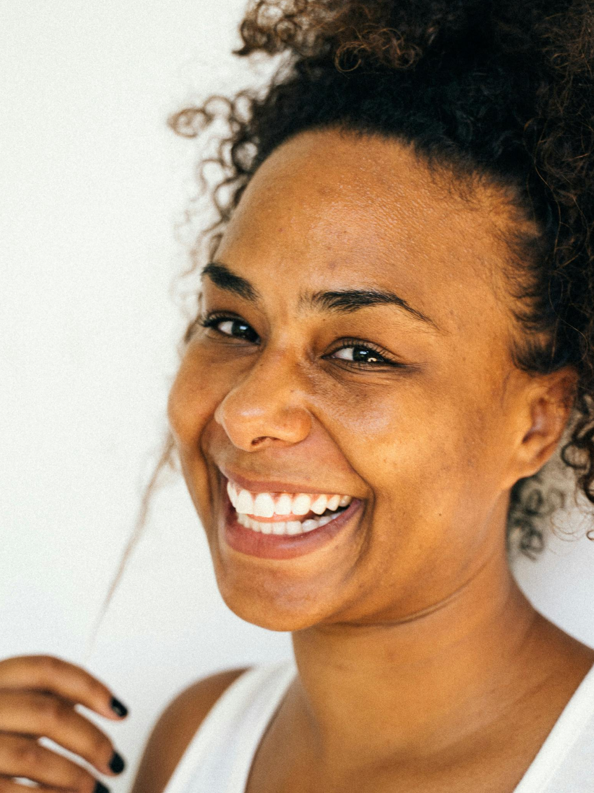 A person with curly hair smiles brightly, wearing a white top and gently touching their hair, reminiscent of an elegant Elementor design against a plain background.