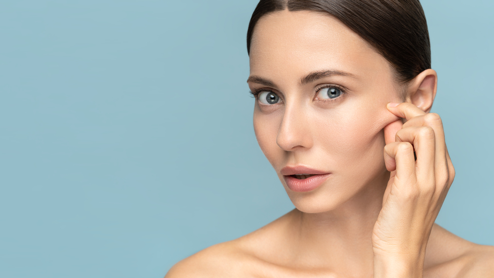 Woman without makeup touching cheeks, has signs of aging skin on her face, looking at camera