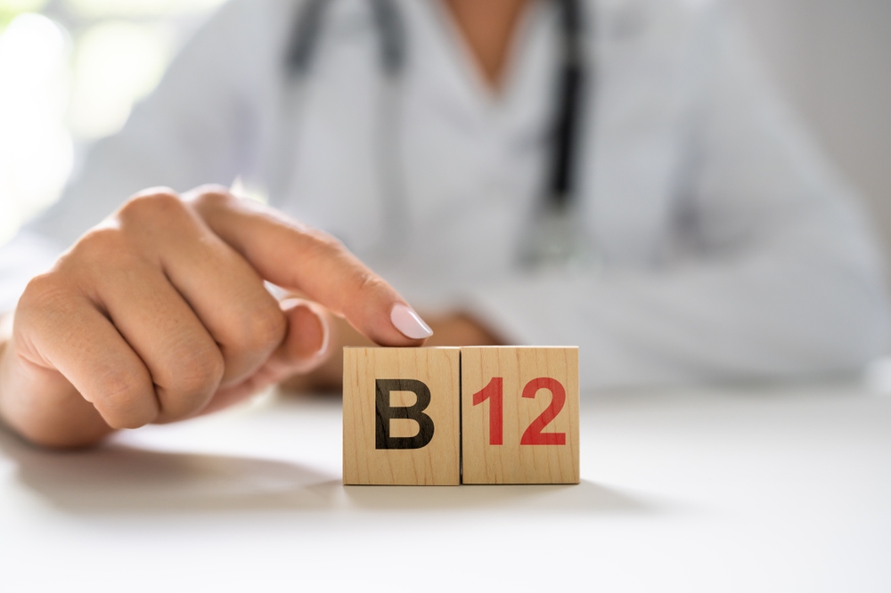 A person wearing a white coat points to wooden blocks displaying "B" and "12" on a table, illustrating the importance of Vitamin B-12