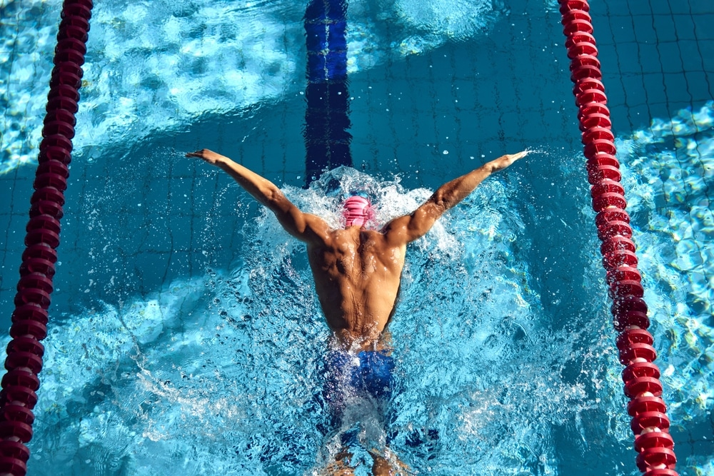 Swimmer, seen from above, powers through water with butterfly stroke, his muscular back glistening under sun as they swim between red lane dividers