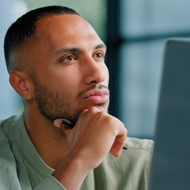 A man with short hair and a trimmed beard holds a pen to his chin and looks thoughtfully at a computer screen, researching the benefits of NAD+ injections.