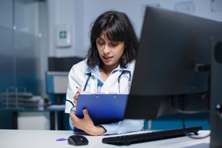 A doctor wearing a white coat sits at a desk, writing on a blue clipboard beside a computer monitor in a medical office, preparing to review COMPREHENSIVE BLOOD PANEL results.